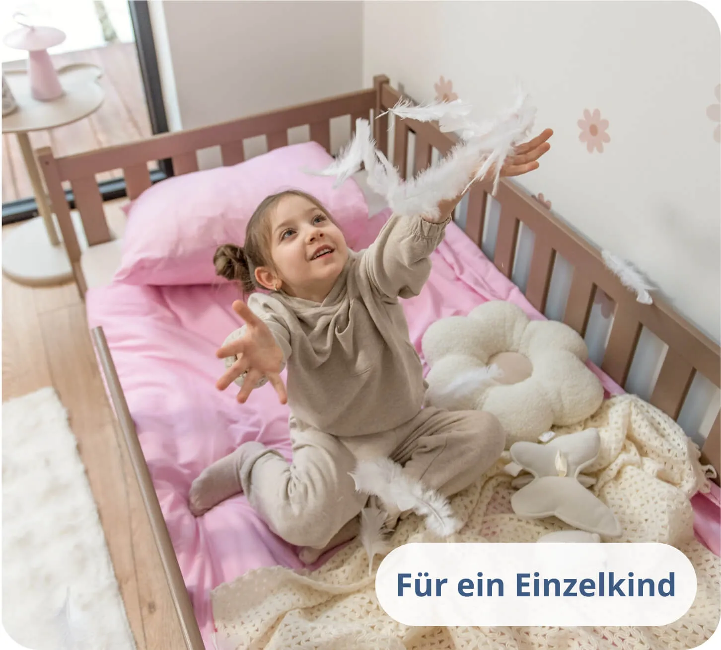 Mädchen spielt mit weißen Federn auf rosa Bettwäsche in einem schokoladenfarbenen Bett.